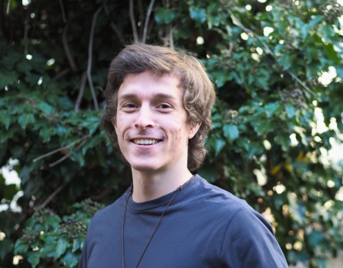 Porträt von Niklas Gentner. Er trägt einen blauen Pulli, hat braune Haare und steht lächelnd vor einem Efeu-Gebüsch.